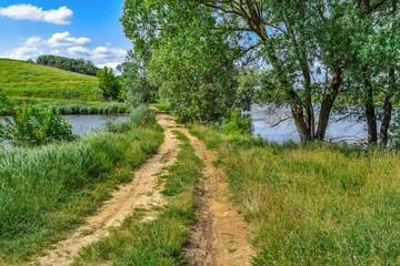 Fototapeta na wymiar Sand-clay two-lane dirt road between two ponds. Beautiful natural landscape with dam among forest lakes and bright young greenery on a sunny spring-summer day (Uhroidy village, Sumy region, Ukraine)