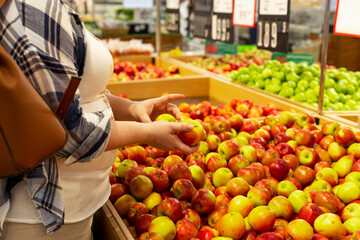 Young woman in a supermarket. Woman holding red apples while choosing fresh fruits and vegetables at farmers market, copy space
