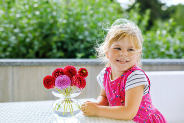 Portrait of little toddler girl admiring bouquet of blooming red and pink dahlia flowers. Cute happy child smelling and counting flower on sunny summer day.