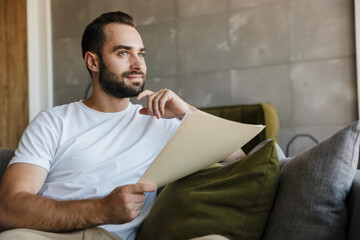 Fototapeta premium Image of young thinking man working with papers while sitting on couch