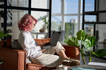 Stylish creative hipster young woman, teen girl with pink hair using pc laptop computer browsing...
