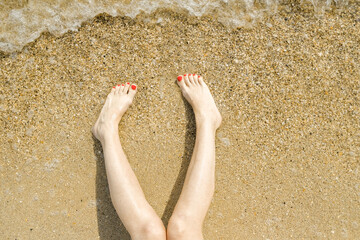 Top view of beautiful female feet with bright red pedicure on the sand of the beach. The sea wave washes women's feet. Relaxation and enjoyment during your seaside holiday. Copy space