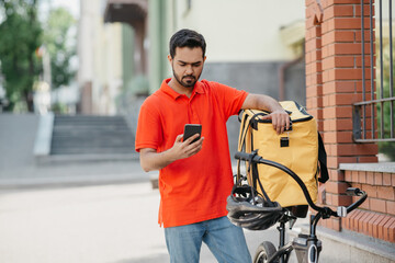 Eco transport and fast food delivery in city. Young cyclist with bag on street, looking customer address