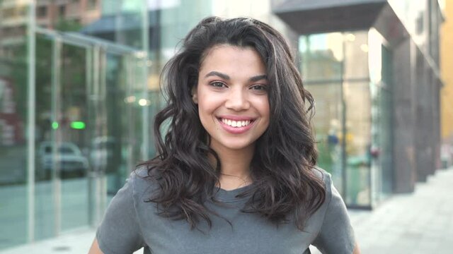 Happy stylish young mixed race gen z girl wearing tshirt standing on city street looking at camera, portrait. Pretty millennial Afro American ethnic woman dental smile closeup outdoor face headshot.