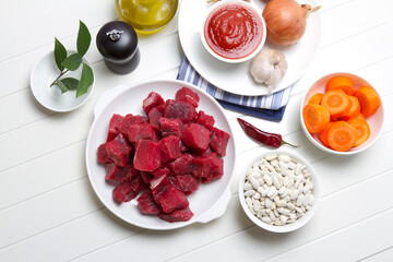 Raw beef, white beans, onions, carrots, garlic, bay leaf, pepper, tomato paste and olive oil on a white table
