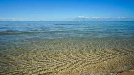 View across calm blue sea to clouds on the horizon, with a gentle wave of clear water washing over tidal ripples on the sandy shore. Burrum Heads, Queensland, Australia.