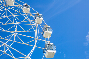 Big ferris wheel with white cabins in an amusement park against a blue sky during the sunny day
