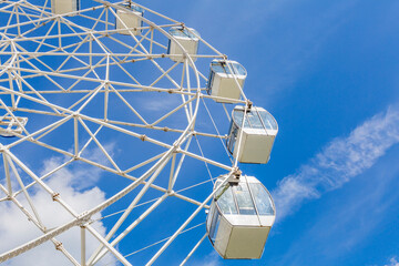 Big ferris wheel with white cabins in an amusement park against a blue sky during the sunny day