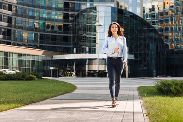 A young business woman in a shirt walks through the business part of the city with a laptop in her hand against the background of a business center, full-length photo