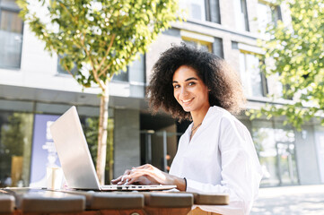 Beautiful African-American businesswoman using laptop computer for work remotely outdoors. A smiling girl in white shirt sits at the table in municipal park with a laptop