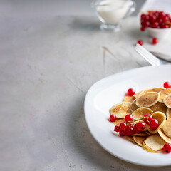 Tiny pancake cereal on white plate served with red currant berries and ingredients on grey cement table. Trendy breakfast, small round baked crepe batter drops. Homemade food. Square with copy space