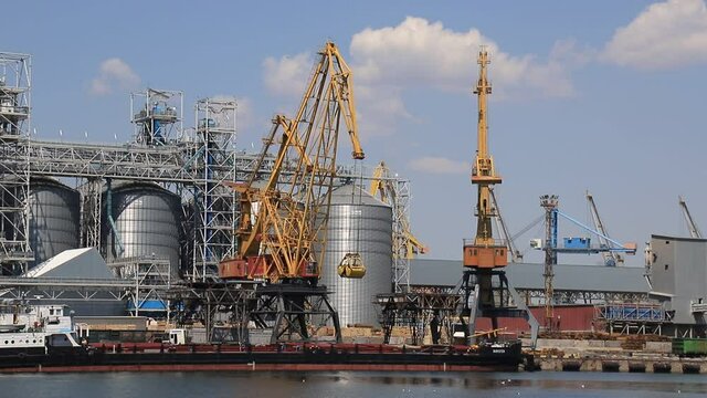Odesa, Ukraine. Yellow harbor cranes unload a barge at the Odessa International Seaport amid large metal tanks. Big cargo port at sea, delivery, unloading of ships, storage of goods