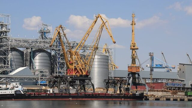 Odesa, Ukraine. Yellow harbor cranes unload a barge at the Odessa International Seaport amid large metal tanks. Big cargo port at sea, delivery, unloading of ships, storage of goods