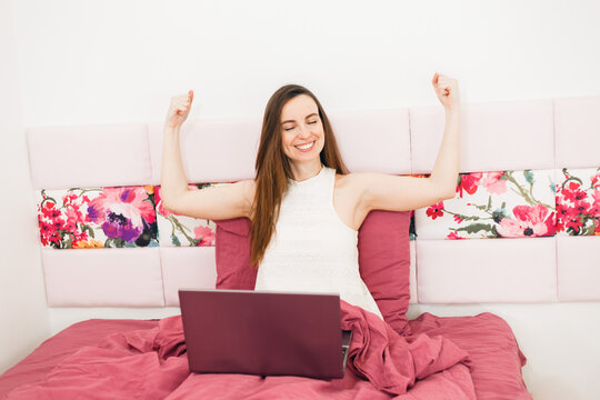 Caucasian young woman working at home. She is using laptop, sitting in bed, raise hads up. Winning jesture