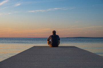 Man in hoodie and short cargo pants sitting on the edge od dock and meditating. Sea horizon, island...