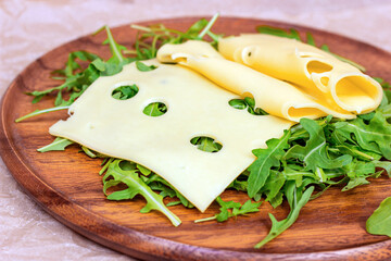 Fresh yellow swiss cheese rolled slices on green arugula leaves on wooden background in the kitchen.
