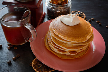 Still life on dark background. Pancakes with sour cream in pink plate, coffee in glass cup with spoon, vintage crusher, dried lemon slices. Close-up.