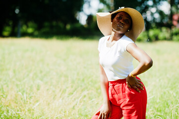 Portrait of gorgeous african american woman 20s wear in summer hat, red pants and white t-shirt, posing at green grass in park.