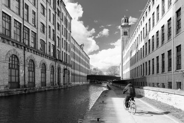 A man is cycling by a canal between historic textile mills in Shipley, UK.