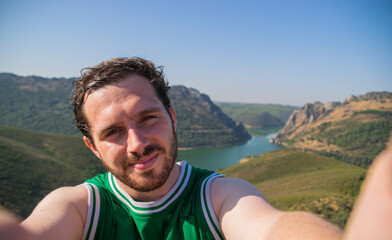 Naklejka premium Photo of a young and attractive guy with beardtaking a selfie while travelling with a landscape behind him. Happy expression. Nature tourism