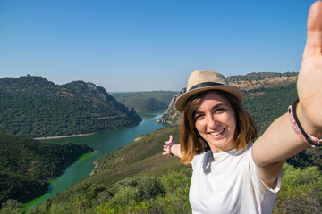 Photo of a young and attractive girl taking a selfie while travelling with a landscape behind her. Happy expression. Nature tourism