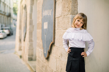 Front view of little blond girl in black and white clothes posing at the street city against the background of the wall of old building