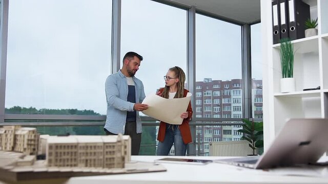 Good-looking contented creative young bearded man and woman with dreadlocks standing near the big window in design office and discussing blueprint of new buildings