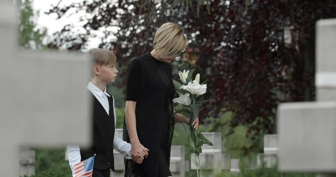 Young widow with flower holding her son hand while walking. Teen kid with american flag looking at his mother while going to honour his father at cemetery Concept of memorial day