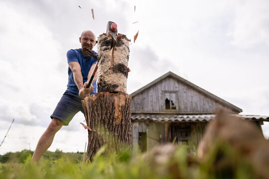 Strong man lumberjack choping wood for the winter, with a large steel ax, in the village, against the background of an old barn.