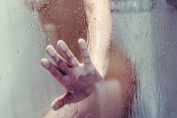 Naked beautiful woman taking a shower and touches hand wet misted glass.