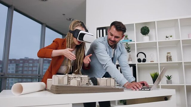 Portrait of happy smiling experienced modern young architects which working together with wooden model of future buildings in design office using information from laptop and augmented reality glasses