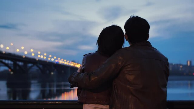 Rear view of a young couple hugging in the evening against a background of blured city lights. Lovers stand on the embankment and watch the evening city. The lights are reflected in the water. Slow