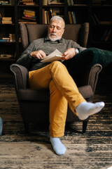 Portrait of handsome bearded mature man sitting on chair and reading book at cozy living room. Happy gray-haired senior male enjoying reading at home.