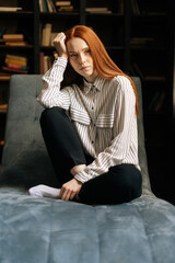 Obraz premium Sad attractive red-haired young woman sitting in chair and looking at the camera on background of home interior. Depressed pretty lady feeling upset while spending time at home alone.