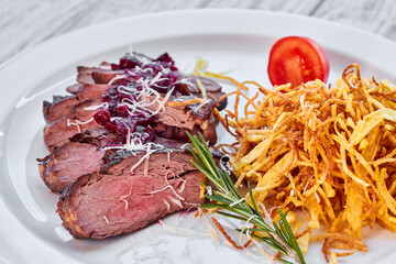 Sliced baked beef with french fries, sauce, tomatoes and rosemary on a white plate and a light wooden background