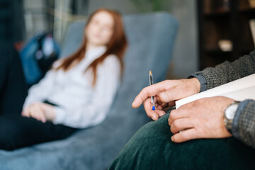 Close-up view of hands of mature man psychologist writing on clipboard while listening to depressed red-haired young woman patient during counseling therapy. Concept of psychological treatment.