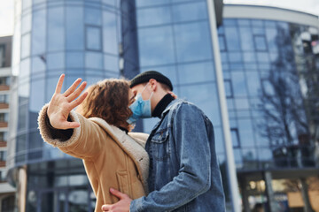 Couple in protective masks kissing each other in the city near business building at quarantine time. Conception of coronavirus