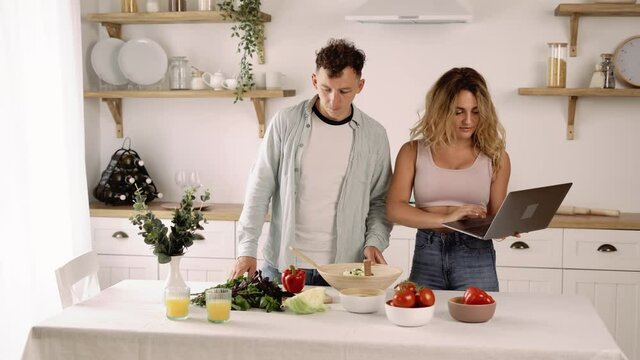 Young Joyful Couple Cooking Healthy Vegetable Salad, Checking or Searching Video Recipe Online on the Laptop, Laughing and Having Fun in the Kitchen. Husband and Wife Preparing Vegeterian Meal Using