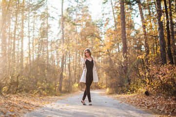 A beautiful, sexy girl in a raincoat stands on the road in the autumn forest at sunset. Warm light. Yellow foliage