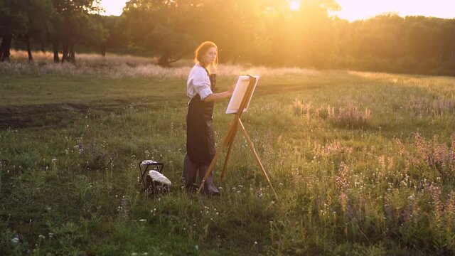 a girl artist with red hair in a beret and a white shirt with an apron draws a landscape on an easel in the evening at sunset