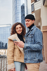 Using phone. Cheerful couple in casual warm clothes have a walk outdoors in the city near business building