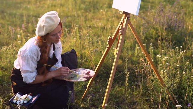 a girl artist with red hair in a beret and a white shirt with an apron draws a landscape on an easel in the evening at sunset