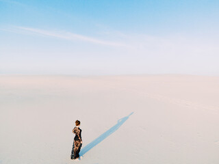 Woman walks in desert and casts a shadow on white sand.