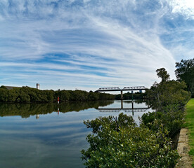 Beautiful view of a river with reflections of a tall pedestrian and water pipe  bridge, trees, deep blue sky and puffy clouds on water, Parramatta river, Rydalmere, Sydney, New South Wales, Australia
