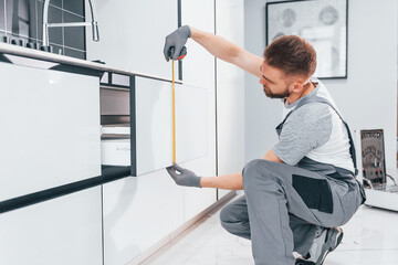 Young professional plumber in grey uniform measuring table by using meter on the kitchen