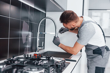 Young professional plumber in grey uniform fixing water tap on the kitchen