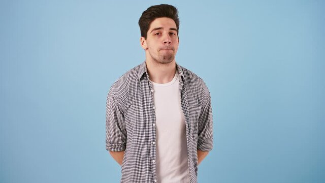 Young handsome male in casual outfit is looking around and showing be quiet sign while posing on blue studio background. Close up