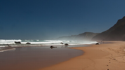 Ocean coast with footprints on sandy beach, black rocks, blue sky and huge waves. Portugal, west coast