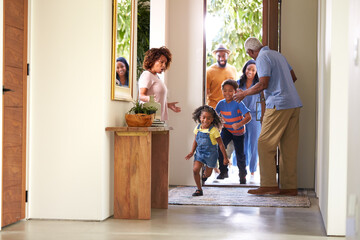 Grandparents At Home Opening Door To Visiting Family With Children Running Ahead