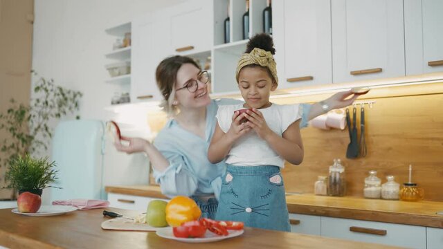 Young white woman in playful mood holds round slices of an apple in her hands and applies them to face of beautiful little African girl depicting glasses. Family is having fun together in kitchen.